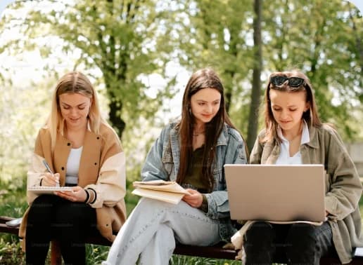 Three students working together with notebooks and a laptop on a bench outdoors